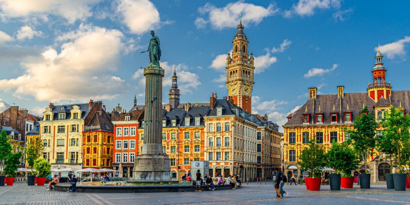 Vue panoramique de la Grand-Place de Lille avec son architecture colorée, la colonne de la Déesse et le Beffroi sous un ciel bleu.